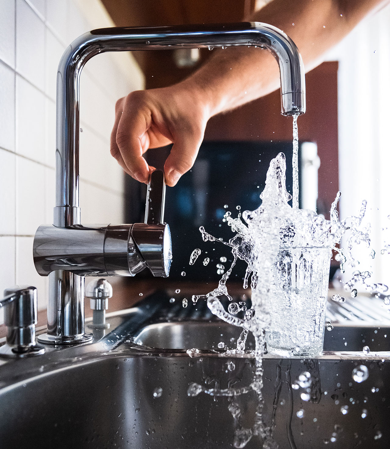 Nærbilde av kjøkkenglass som blir fylt med vann så det spruter over, fra blandebatteri på kjøkkenbenk i stål og tre.